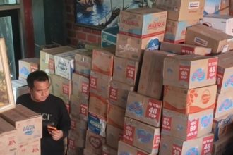 Cak Sholeh berada di tengah tumpukan duz air mineral dalam kemasan hasil donasi masyarakat. (foto: jero)